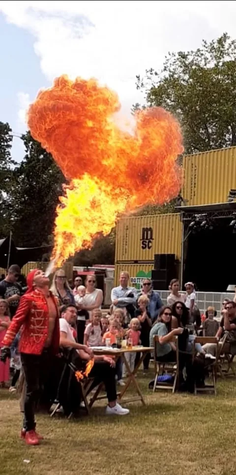 Vuurspuwer Nuno in rood circusjasje blaast een vuurbal boven het publiek op een zomers familiefestival