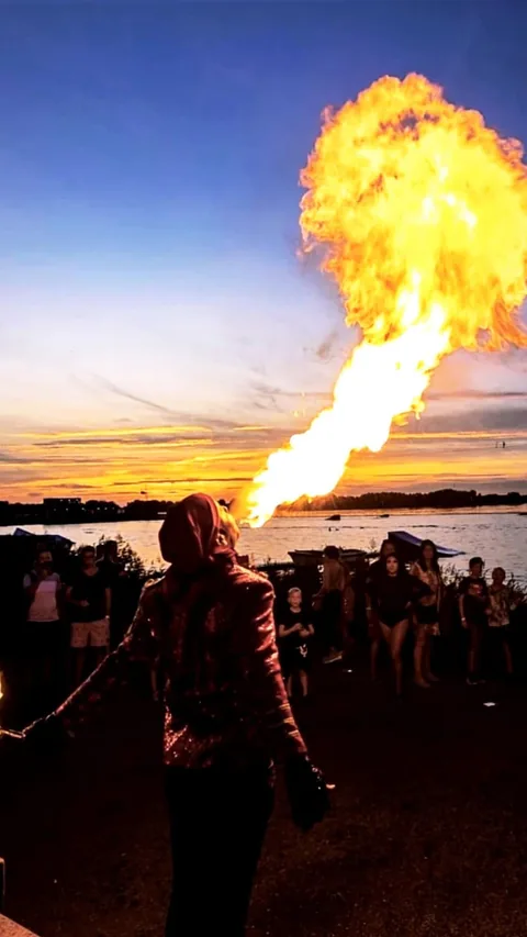 Vuurspuwer Nuno in glitterjasje blaast een vuurbal over het water bij zonsondergang voor publiek